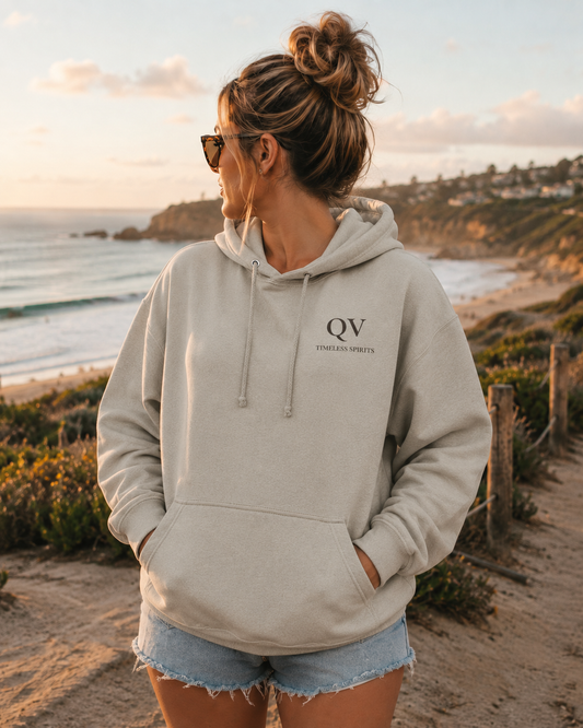 Woman wearing the Queer Vintage Timeless Spirits ash grey hoodie at sunset overlooking the coast, featuring the minimalist QV logo on the chest