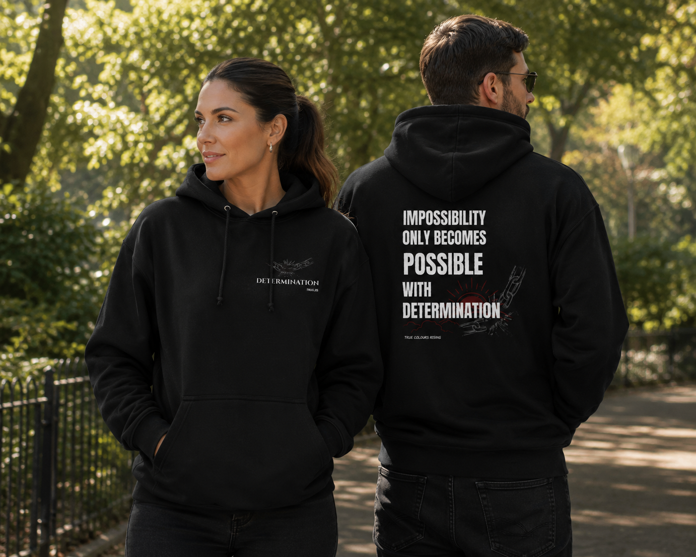 Couple wearing matching IMPOSSIBILITY BECOMES POSSIBLE WITH DETERMINATION jet black hoodies in a leafy park, showing both the front chest motif and bold back graphic.