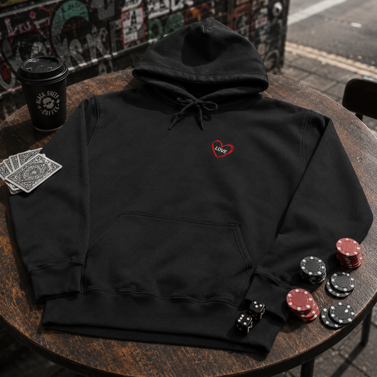 Folded Love Is a Gamble jet black hoodie displaying the gothic tattoo-inspired artwork beside casual jeans and trainers.
