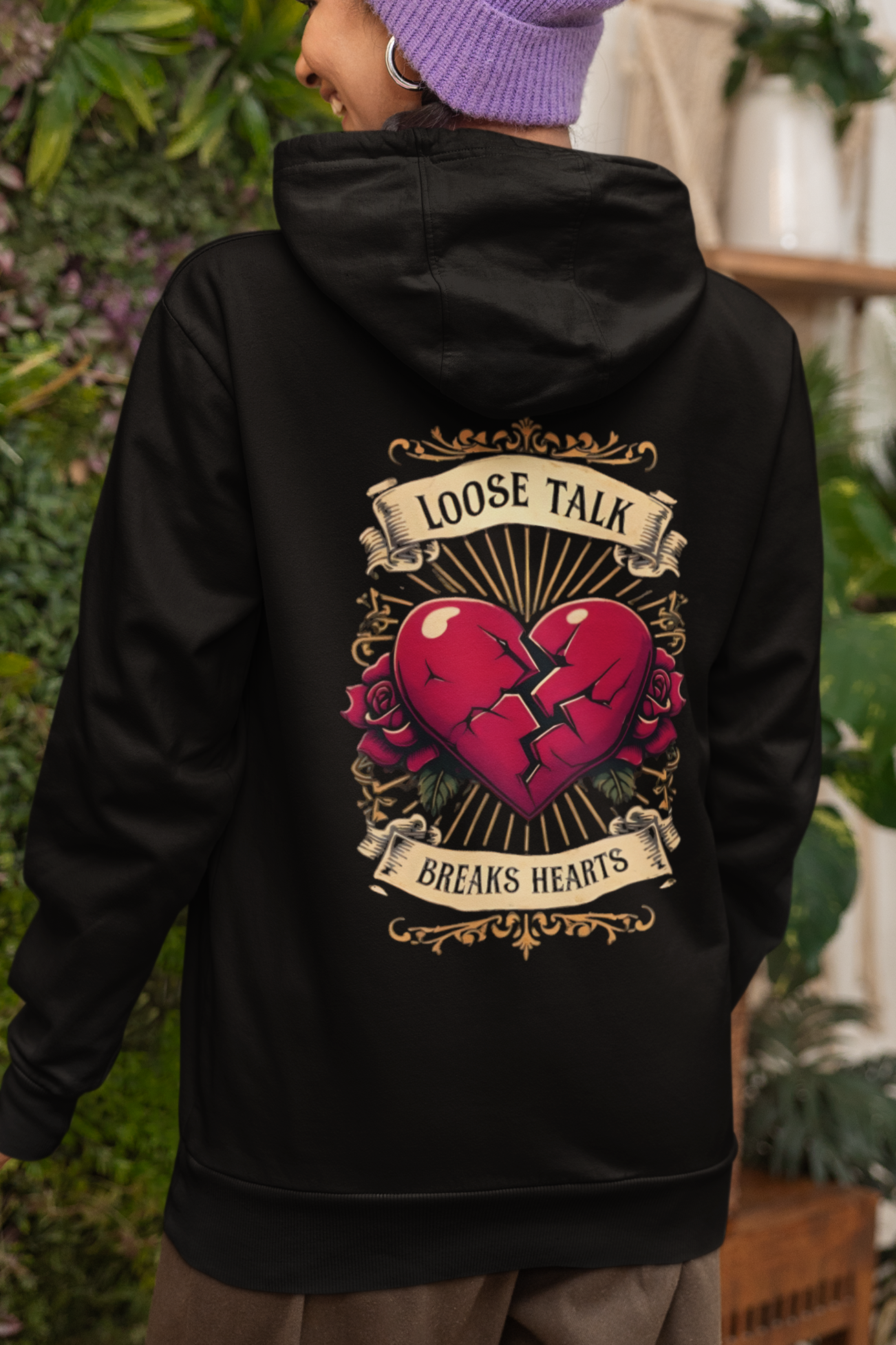 Person wearing a black hoodie with a vintage graphic design on the back, standing in an indoor setting with plants. heart broken but healing