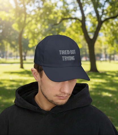 Angled view of a navy unstructured dad cap featuring minimalist “Tired But Trying” embroidered lettering