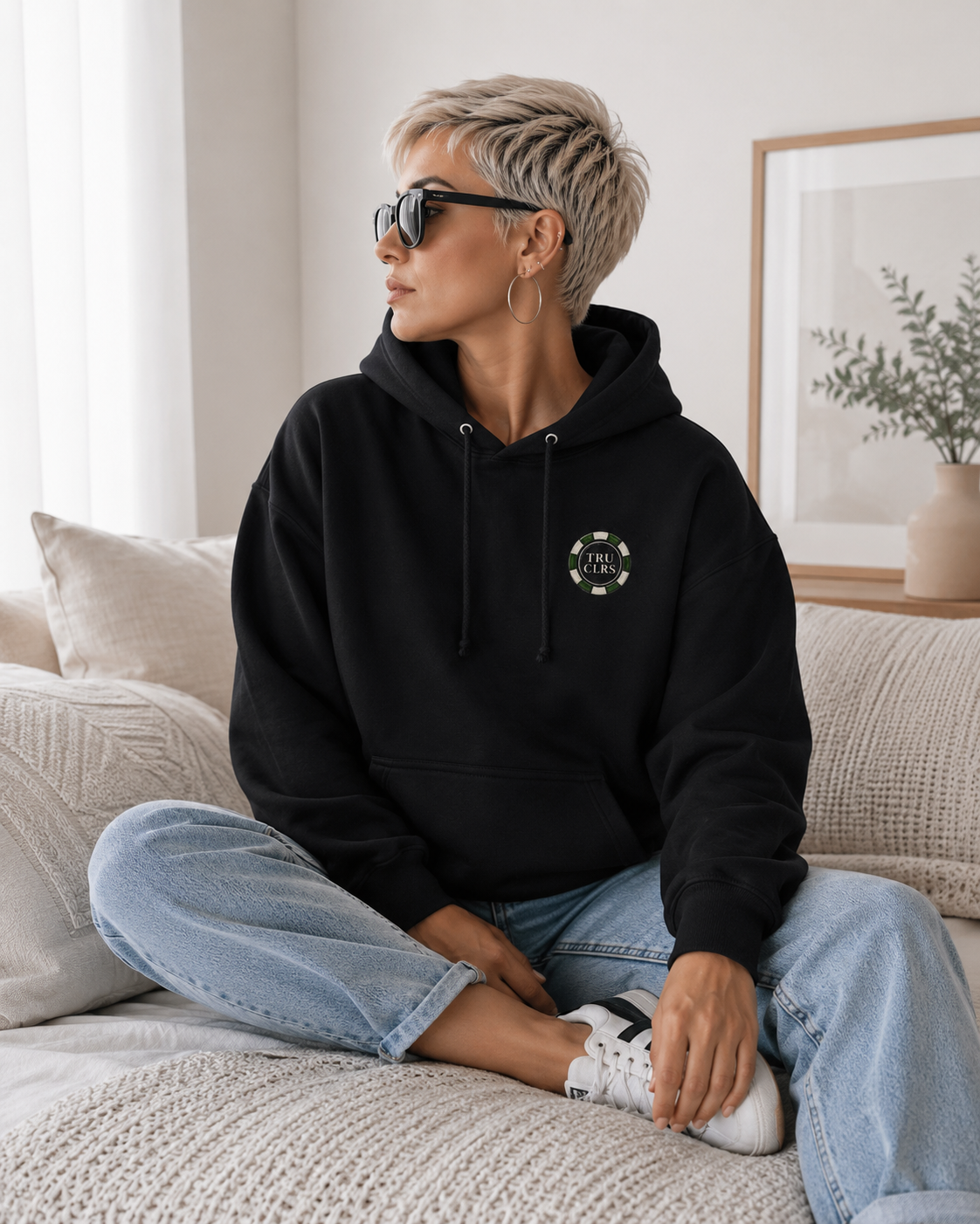 Lucky Shot black hoodie with TRU CLRS chest logo styled on a light sofa with blue jeans and white trainers in a bright modern lifestyle setting.