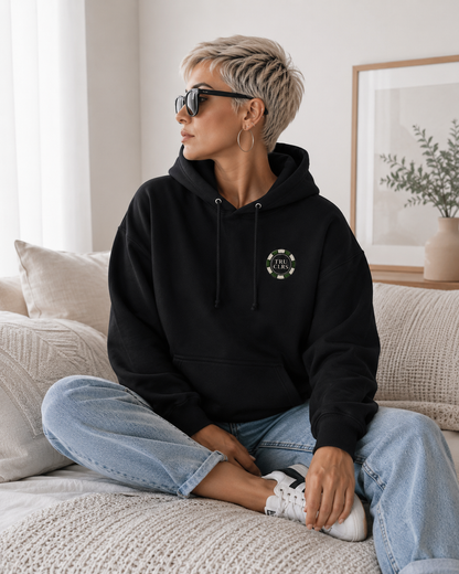 Lucky Shot black hoodie with TRU CLRS chest logo styled on a light sofa with blue jeans and white trainers in a bright modern lifestyle setting.