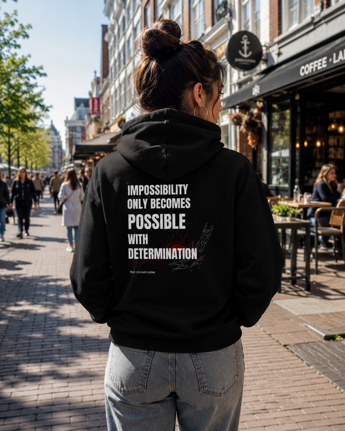 Woman wearing the IMPOSSIBILITY BECOMES POSSIBLE WITH DETERMINATION black hoodie from the back on a sunny city street with cafés, showcasing the large statement print.