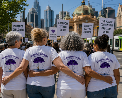 Four women wearing shirts that say 'Grandmothers of The Hauged -SHOUT. SHELTER. SUPPORT.' holding hands at a protest.